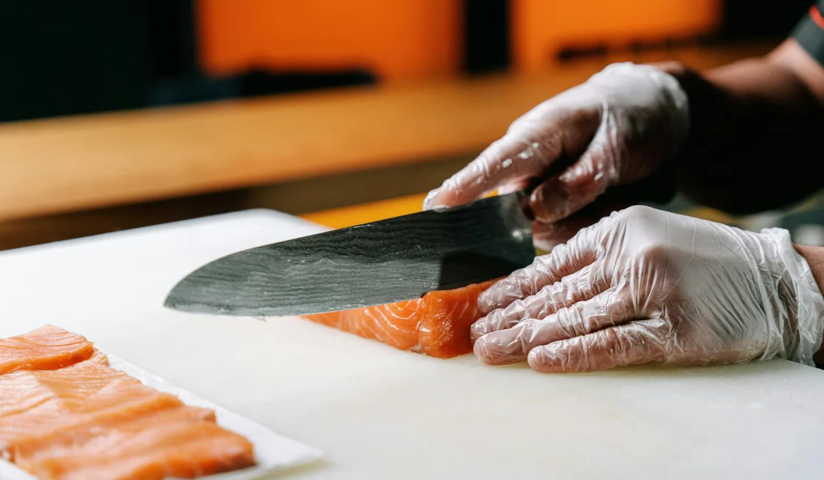 Gloved hands slicing a salmon fillet on a white cutting board with a knife