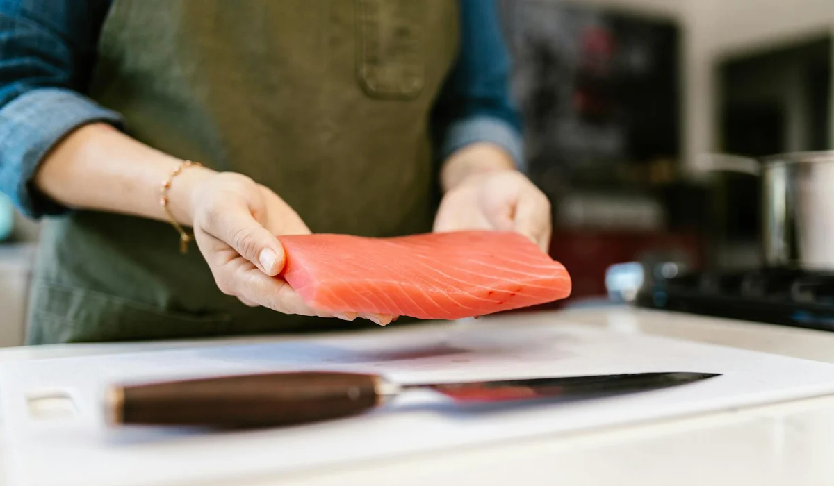 A person wearing a green apron holds a raw salmon fillet over a white cutting board in a kitchen, with a knife resting nearby.