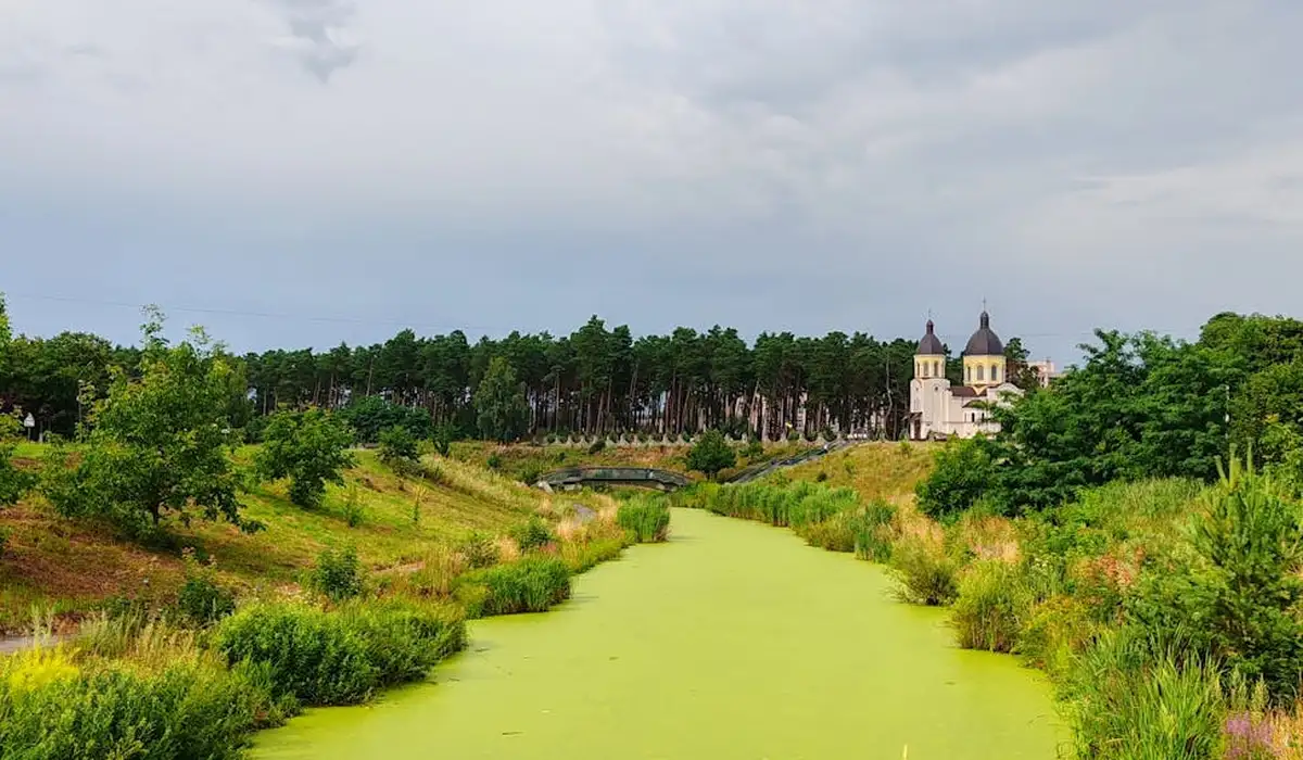 Algae-covered waterway winding through a grassy landscape with a distant building and a cloudy sky.