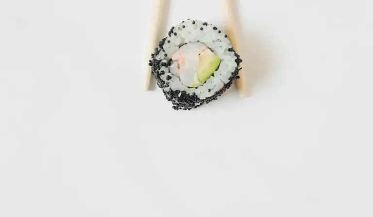 Sushi roll with avocado filling and sesame seeds, held by chopsticks on a white background
