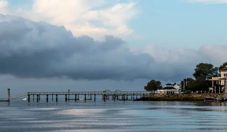Calm coastal water with a wooden pier extending into the harbor and a cloudy sky above.