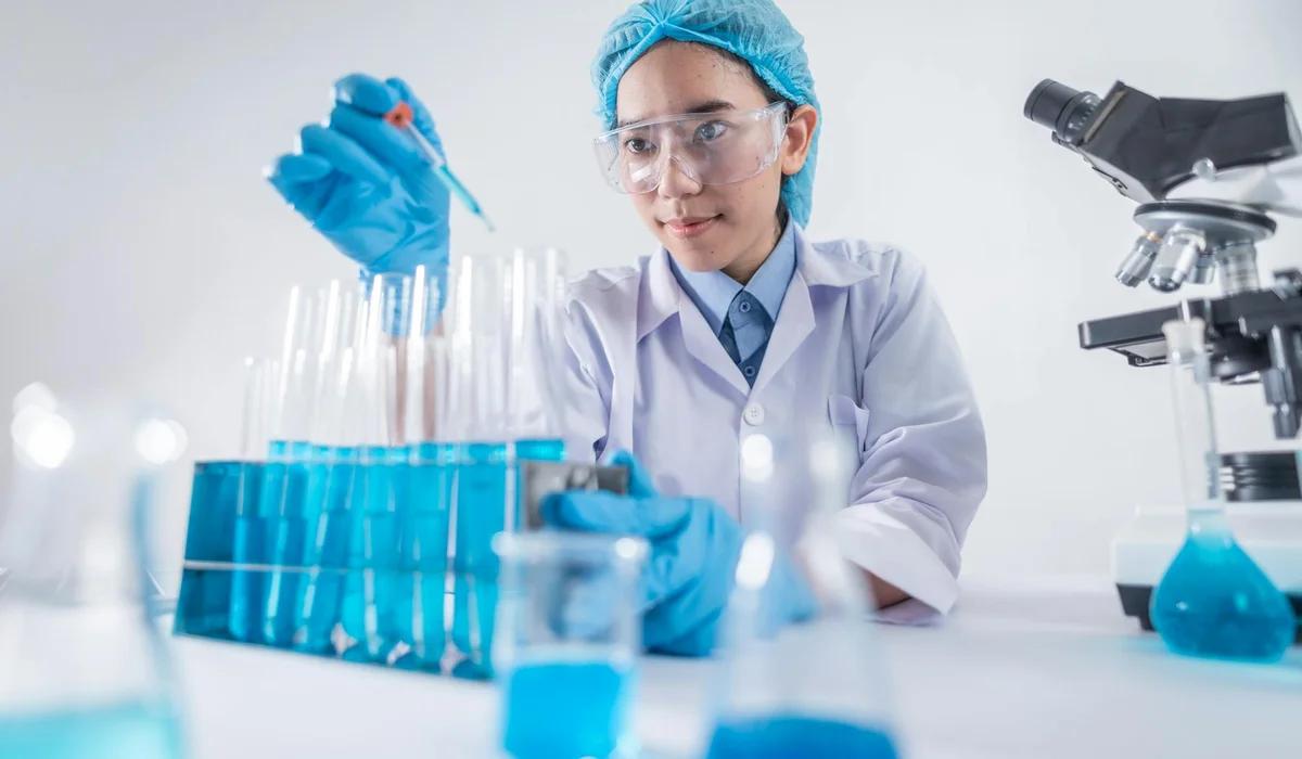 A lab scientist in safety gear, wearing gloves and a hairnet, uses a pipette to transfer blue liquid between test tubes, with a microscope and other lab glassware visible on the bench.